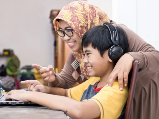 A mother helping her young son homeschool. The son is sitting at his desk with his laptop and headphones on, and they are both smiling and pointing at the screen.