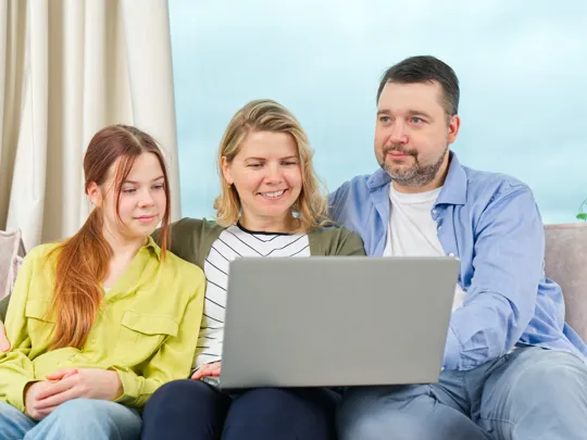Family of three sitting together on a sofa, smiling while viewing a laptop screen.