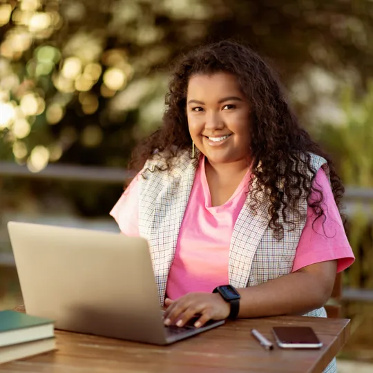A smiling young student with long, curly dark hair sits outdoors at a wooden table, working on a laptop. She is wearing a bright pink shirt and a light vest. A stack of books, a smartphone, and a pen are on the table next to her, with a blurred natural green background.