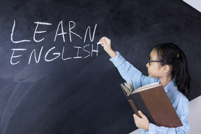 Young student writing “Learn English” on a blackboard while holding a book in one hand
