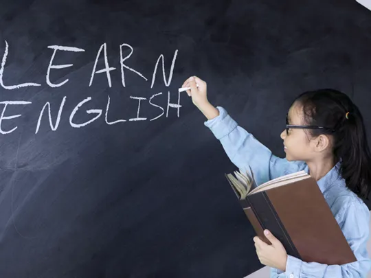 Young student writing “Learn English” on a blackboard while holding a book in one hand
