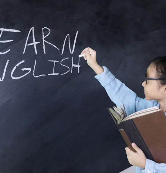 Young student writing “Learn English” on a blackboard while holding a book in one hand