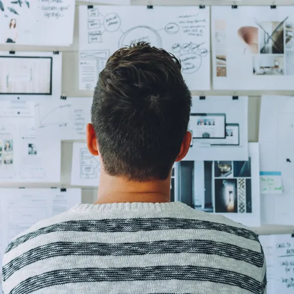 A person is seen from behind, standing in front of a wall covered in papers, sketches, and notes. The person is wearing a striped sweater.