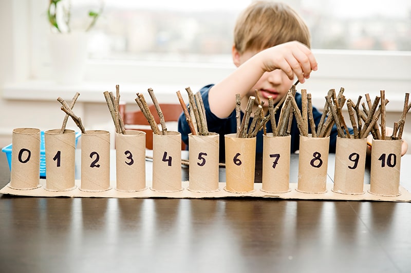 Child placing sticks into cardboard tubes labelled 0 to 10, exploring number recognition and counting