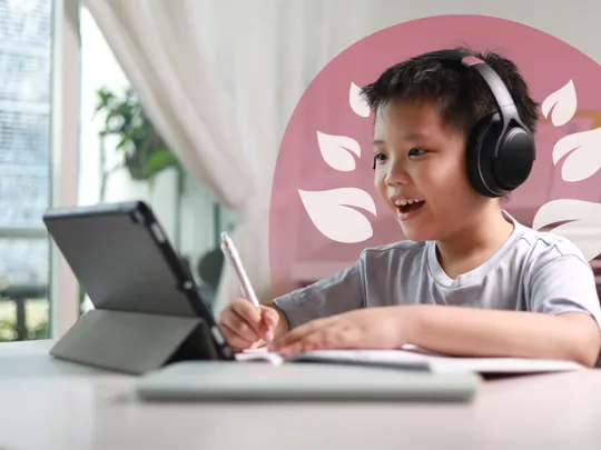 A young student wearing headphones, smiling and writing in a notebook while looking at a tablet, indicating active online learning.
