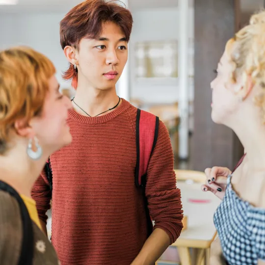 Three young university or high school students are engaged in a conversation indoors, possibly in a classroom or cafe setting. A male student in a terracotta-colored sweater is looking intently at a female student with curly blonde hair. Another female student is visible on the left.