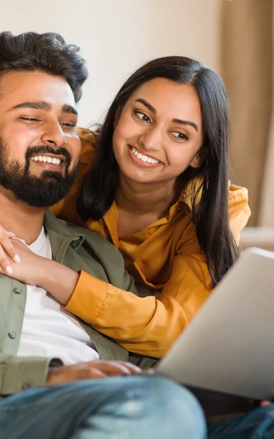 Couple smiling warmly while looking at a laptop together on a cosy living room sofa.