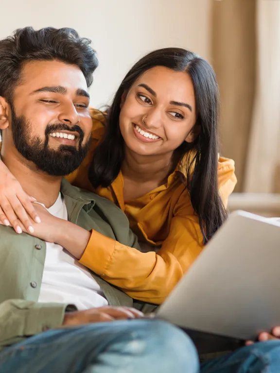 Couple smiling warmly while looking at a laptop together on a cosy living room sofa.