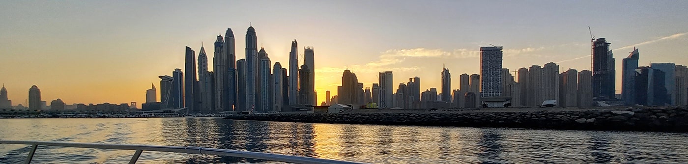 A panoramic view of a city skyline at sunset, with tall buildings reflecting in the water of a harbour or bay.