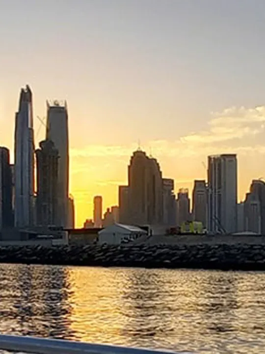 A panoramic view of a city skyline at sunset, with tall buildings reflecting in the water of a harbour or bay.