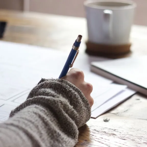 A close-up, over-the-shoulder shot of a person's hand holding a pen and writing on a piece of paper. The person is wearing a gray sweater. A coffee cup and a notebook are on the wooden table.