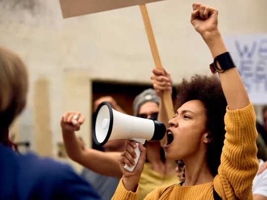 Woman raising her fist and speaking into a megaphone during a public protest.