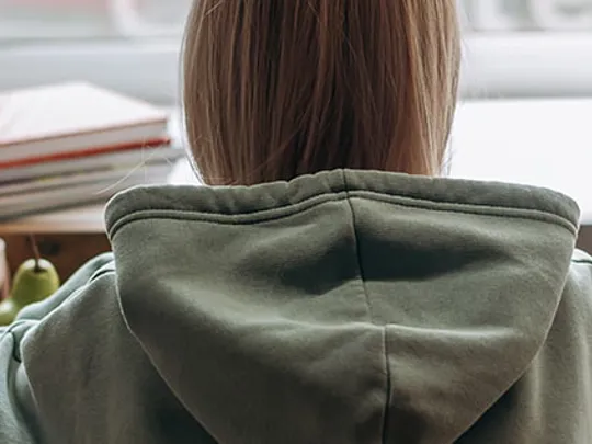 The back of a student wearing a grey hoodie, sitting at a desk with books, a globe, and stationery, looking towards a window.