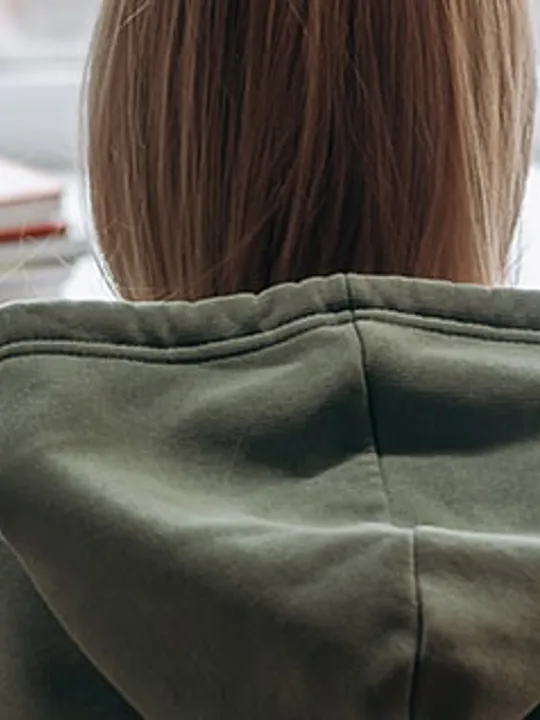 The back of a student wearing a grey hoodie, sitting at a desk with books, a globe, and stationery, looking towards a window.