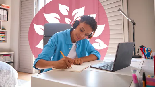 A young person wearing headphones, writing in a notebook at a desk with a laptop and school supplies.