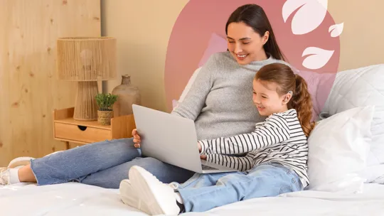 A woman and a child are comfortably seated on a bed, both smiling and looking at a laptop together. The woman is wearing a grey sweater and jeans, while the child is in a striped long-sleeve shirt and jeans. A nightstand with a lamp is visible to the left. A translucent pink overlay with white leaf designs is in the top right.