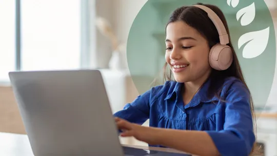 A young student wearing pink headphones, smiling and looking at a laptop while typing.