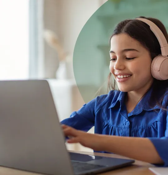 A young student wearing pink headphones, smiling and looking at a laptop while typing.