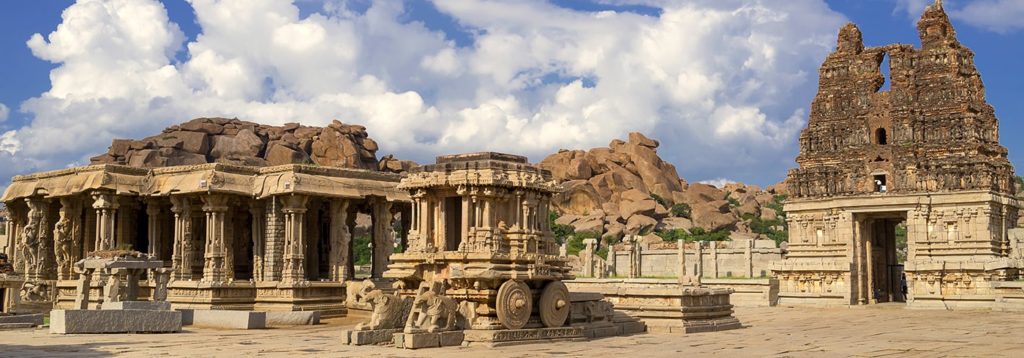 Ornate stone carvings and temples at a historic Indian site with boulder-strewn hills in the background.
