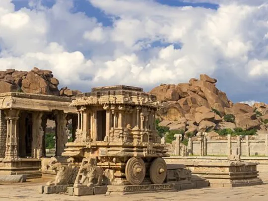 Ornate stone carvings and temples at a historic Indian site with boulder-strewn hills in the background.