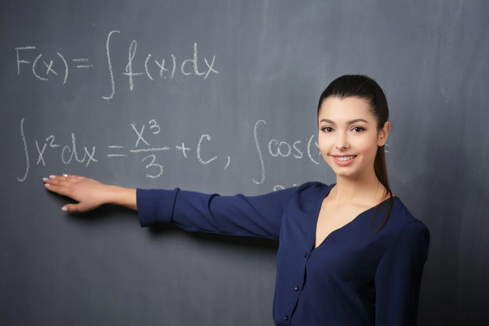 A female maths teacher in front of a blackboard pointing to an equation. 