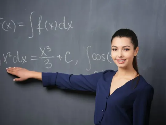 A female maths teacher in front of a blackboard pointing to an equation.