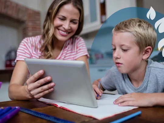 A homeschooling mum and son sitting at a desk. The mum is smiling and showing her son something on a tablet device, which the boy is studying intently.