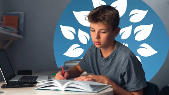 Teen boy studying at a desk, writing in an open book beside a laptop.