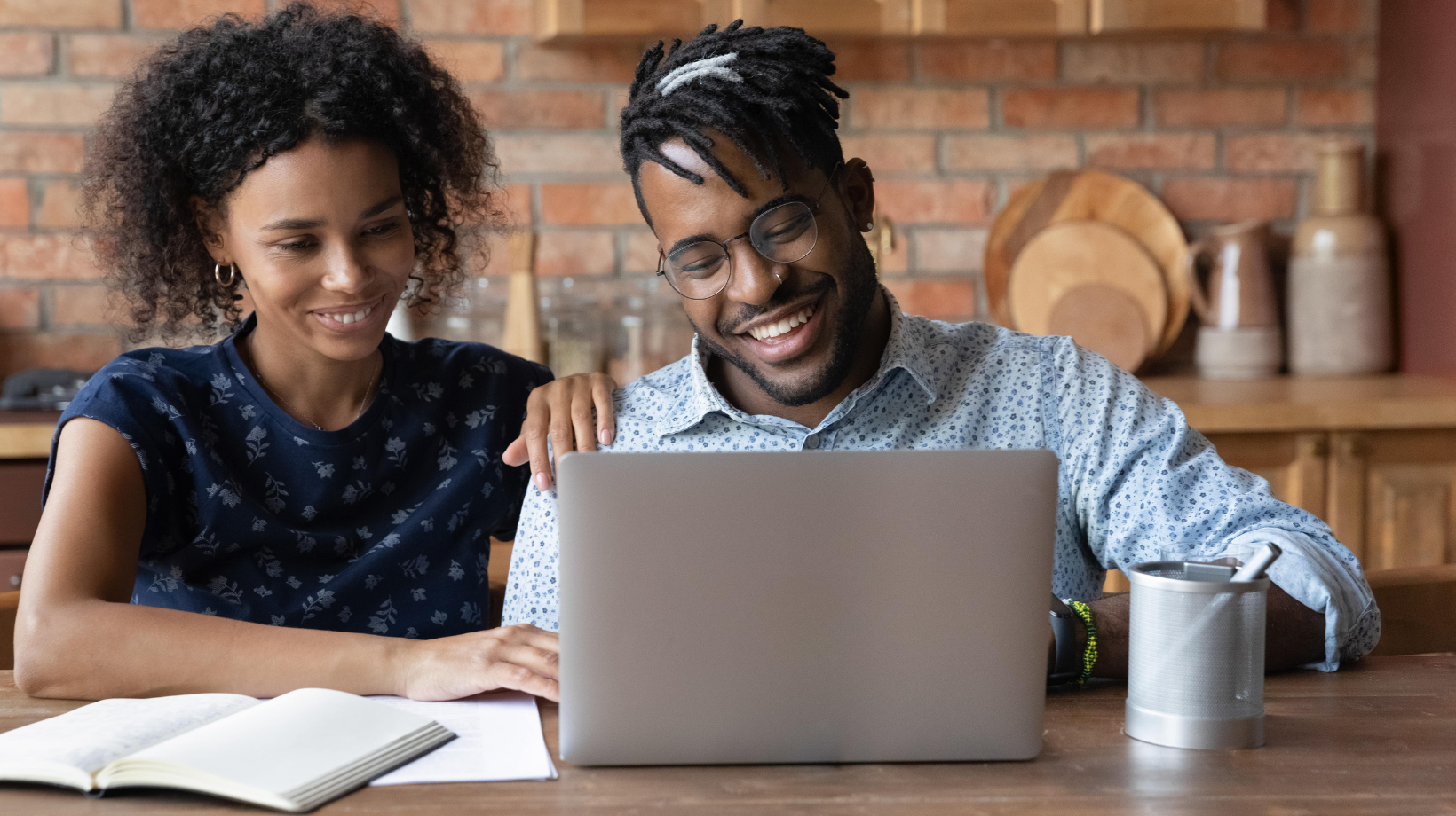 A smiling man and woman sit at a table, using a laptop and a notebook to plan their child's online education together