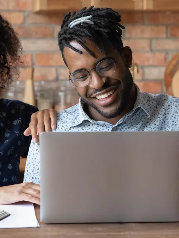 A smiling man and woman sit at a table, using a laptop and a notebook to plan their child's online education together