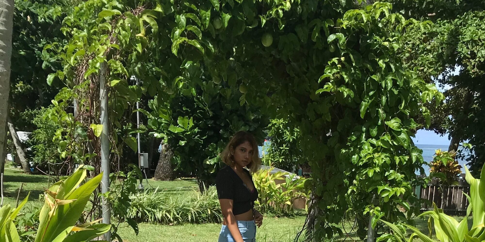 A young woman standing under a green leafy archway with tropical foliage and a glimpse of the ocean in the background in Mauritius.