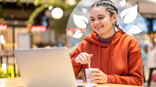 A young person with braided hair smiling while working on a laptop and holding a drink cup in a bright indoor setting.