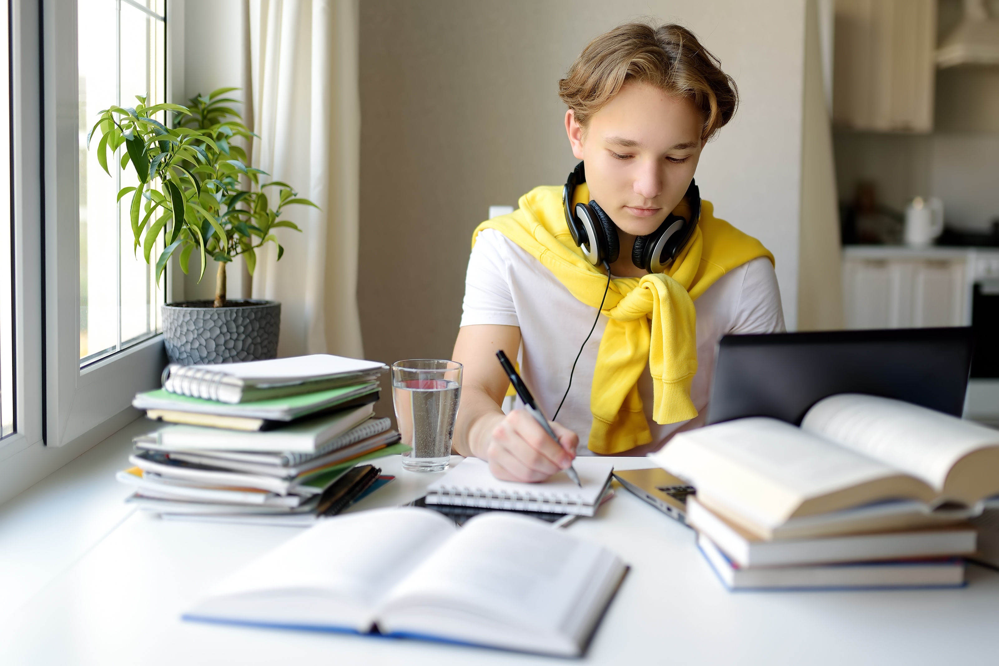A teenage student with headphones sits at a desk at home, writing in a notebook while studying online with a laptop and books.