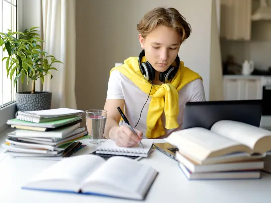 A teenage student with headphones sits at a desk at home, writing in a notebook while studying online with a laptop and books.