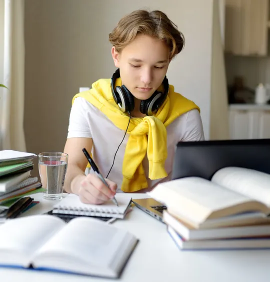 A teenage student with headphones sits at a desk at home, writing in a notebook while studying online with a laptop and books.