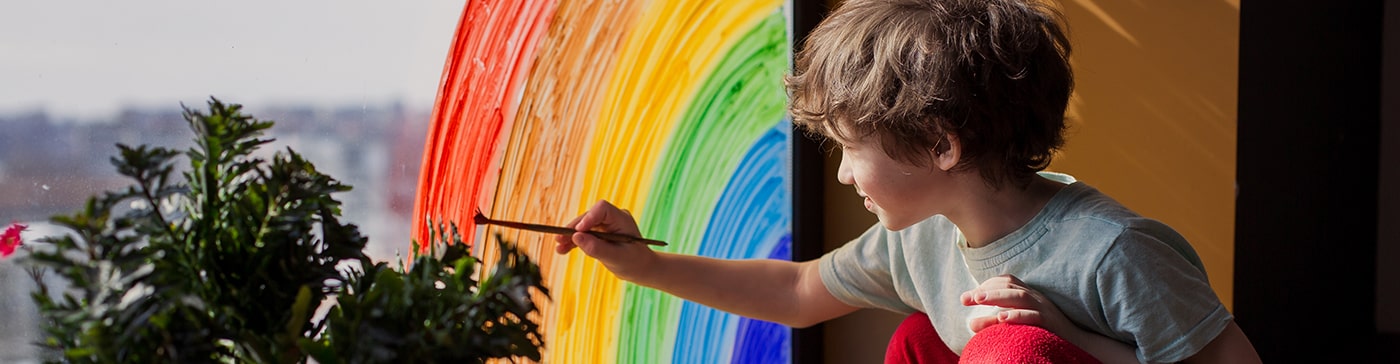 Child painting a rainbow on a window with sunlight streaming in, engaging in a creative art activity