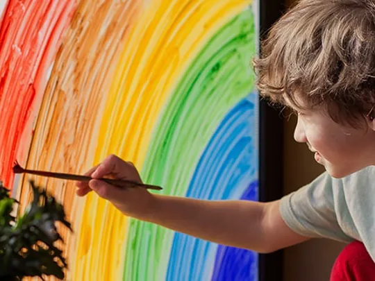 Child painting a rainbow on a window with sunlight streaming in, engaging in a creative art activity