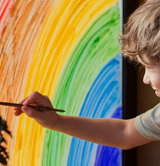 Child painting a rainbow on a window with sunlight streaming in, engaging in a creative art activity