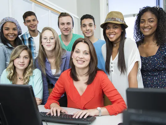 Group of students gathered around their tutor, smiling in a classroom with computers.