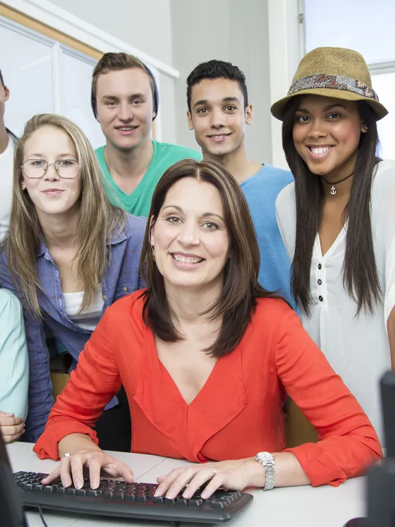 Group of students gathered around their tutor, smiling in a classroom with computers.