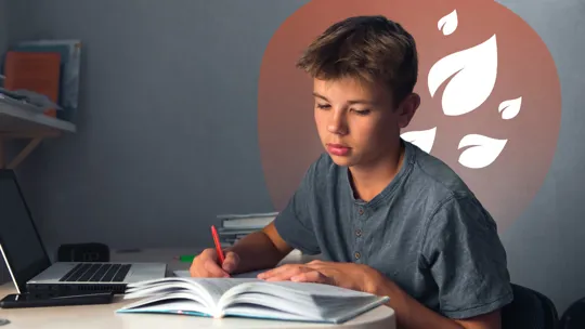 Teen boy studying at a desk, writing in an open book beside a laptop.