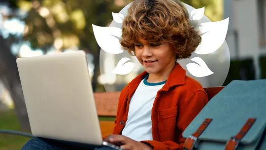 A young student with curly hair, sitting on a bench outdoors and smiling while using a laptop.