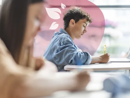 A young student with curly hair is focused on writing during an exam or study session, with a laptop nearby.