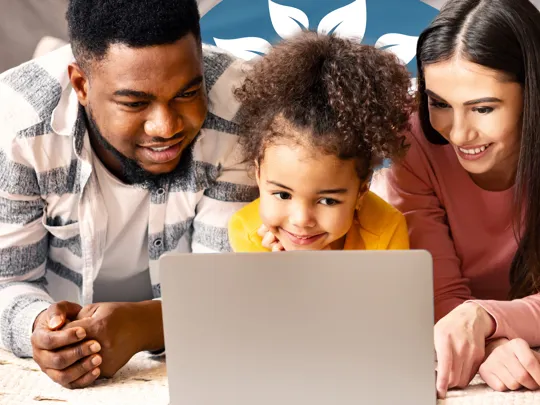 A family lying on the floor, smiling and looking at a laptop together, suggesting engaging online learning or family activities.