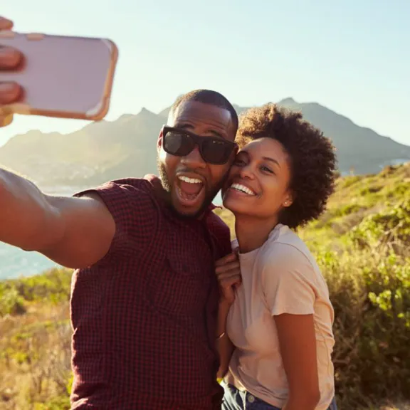 A happy couple taking a selfie with a smartphone while standing on a hill overlooking a body of water and mountains. The man in the foreground is holding the phone and has a wide-open smile, while the woman next to him is also smiling and leaning in.