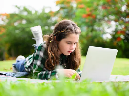 A girl studies comfortably on her laptop in a grassy field, showcasing the flexible learning options available through homeschooling.