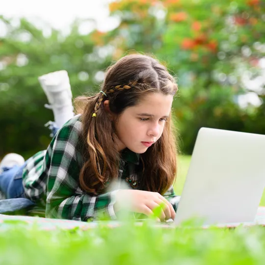 A girl studies comfortably on her laptop in a grassy field, showcasing the flexible learning options available through homeschooling.