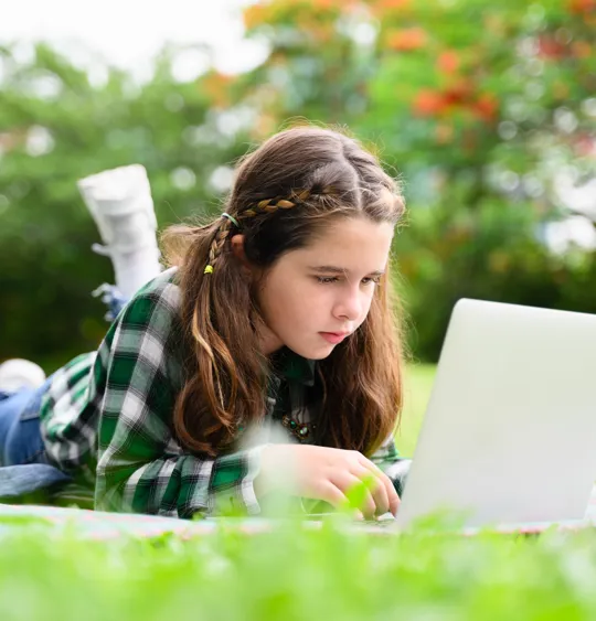A girl studies comfortably on her laptop in a grassy field, showcasing the flexible learning options available through homeschooling.