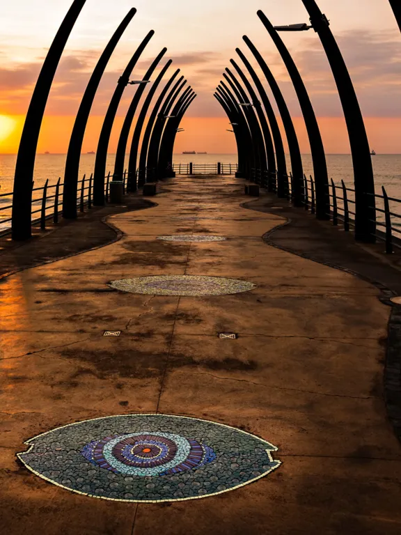 The iconic Whalebone Pier in Umhlanga Rocks, Durban, South Africa, at sunrise or sunset with the sun glowing over the ocean.