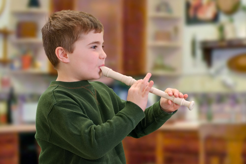 Young boy in a green jumper playing a white recorder in a classroom setting.